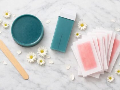 Flat lay con disco de cera caliente azuleno verde azulado, cartucho roll-on del mismo color, espátula de madera y bandas de cera fría rosa con flores de manzanilla sobre mármol blanco