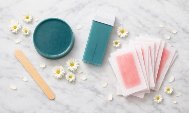 Flat lay con disco de cera caliente azuleno verde azulado, cartucho roll-on del mismo color, espátula de madera y bandas de cera fría rosa con flores de manzanilla sobre mármol blanco