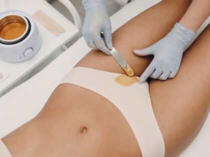 Esteticista con guantes aplicando cera caliente dorada con espátula en la ingle de una mujer tumbada en camilla blanca con calentador de cera al fondo