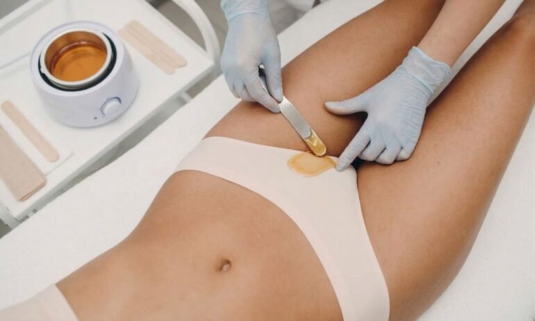 Esteticista con guantes aplicando cera caliente dorada con espátula en la ingle de una mujer tumbada en camilla blanca con calentador de cera al fondo