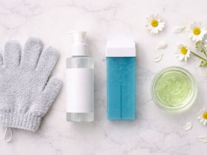 Guante exfoliante gris, botella de loción pre-depil, cartucho roll-on de cera azuleno azul y cuenco de gel de aloe vera verde sobre mármol blanco con flores de manzanilla